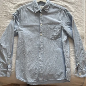 Ljusblå skjorta från H&M - Klassisk ljusblå skjorta från H&M med regular fit. Skjortan har lång ärm, bröstficka och knappar framtill. Perfekt till både jeans och chinos för en clean och enkel stil.