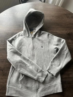 Grå hoodie från Polo Ralph Lauren - Klassisk grå hoodie från Polo Ralph Lauren med dragkedja framtill och två fickor. Köpt på kidsbrandstore december 2025, knappt använd, mycket fint skick. Nypris 1295kr. Storlek 160/L.