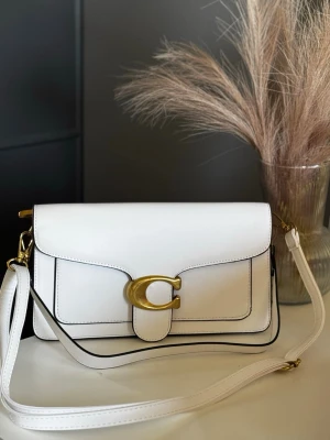 Vit axelväska - Snygg vit axelväska från med justerbar axelrem och guldfärgade metalldetaljer. Väskan har ett stort C-spänne framtill och svarta kanter som ger en stilren kontrast. Perfekt för dig som gillar klassisk men trendig design.