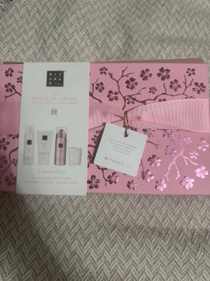 Rituals The Ritual of Sakura presentbox - Lyxig presentbox från Rituals i serien The Ritual of Sakura. Innehåller fyra bästsäljare: shower foam, body cream, body lotion mousse och doftljus. Förpackningen är rosa med glansiga körsbärsblommor och ett matchande band. Perfekt för dig som älskar skönhet och self-care.