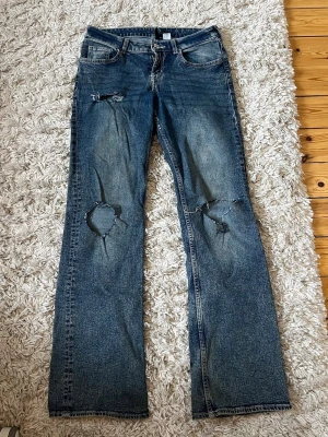 Blå slitna bootcut jeans - Säljer ett par blå bootcut jeans med slitningar och hål på knäna. Jeansen har klassisk femficksmodell, dragkedja och knapp i midjan. Materialet är denim i bomull och passformen är rak med lätt utsvängda ben.