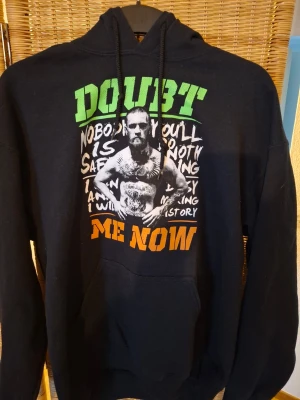 Svart hoodie med tryck och text - Säljer en svart hoodie med stor färgglad text 'DOUBT ME NOW' i grönt och orange samt ett stort tryck av en muskulös person på framsidan. Hoodien har huva med snören och en klassisk magficka. Perfekt för dig som gillar streetwear och statement-plagg.