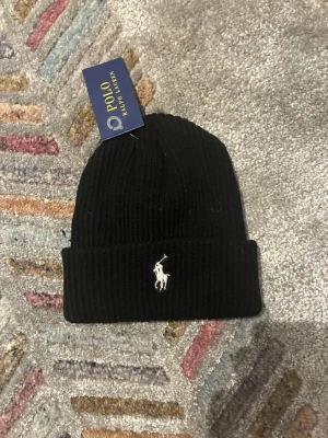 Svart ribbstickad mössa Polo Ralph Lauren - Snygg svart ribbstickad mössa från Polo Ralph Lauren med klassisk vit broderad logga framtill. Mössan har uppvikt kant och är tillverkad i mjukt material som håller dig varm och stilren under kalla dagar.