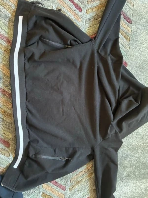 Svart hoodie med vita detaljer - Snygg svart hoodie med dragkedjeförsedda fickor och vita ränder längs ärmslut och nederkant. Hoodien har en klassisk huva och ribbade muddar för extra komfort. Perfekt för en avslappnad streetwear-look.