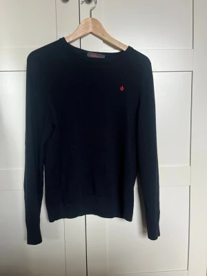 Mörkblå stickad tröja från Morris  - Mörkblå stickad tröja från Morris med rund halsringning och diskret röd logga broderad på bröstet. Tröjan har långa ärmar och ribbade muddar vid ärmslut och nederkant. Perfekt för lager på lager och enkel att matcha.
