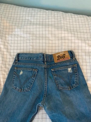 D&G blå raka jeans med slitningar - Ett par blå jeans från D&G i klassisk denim med raka ben och slitna detaljer på bakfickorna. Jeansen har fem fickor, bälteshällor och ett brunt läderpatch med logga bak i midjan. Perfekt för dig som gillar en avslappnad och trendig look. Det finns en liten liten grön färg fläck på bak fickan men den syns knappt