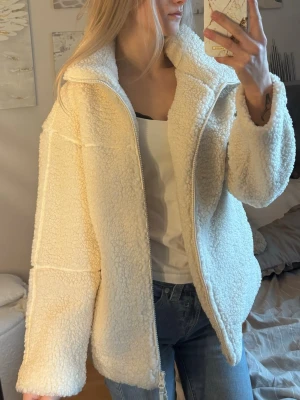 Krämvit teddyjacka med dragkedja - Mysig krämvit teddyjacka med hög krage och dragkedja framtill. Jackan har en oversized passform och är gjord i fluffigt, mjukt teddy-material som ger en avslappnad look. Perfekt att slänga på sig över en hoodie eller t-shirt för extra värme. (Xs men passar S, aldrig använd)