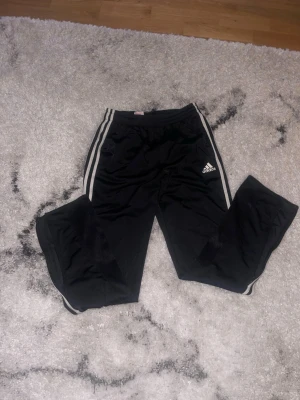 Svarta Adidas träningsbyxor - Säljer ett par klassiska svarta Adidas träningsbyxor med vita ränder längs sidorna och Adidas-logga på vänster ben. Byxorna har resår i midjan och är tillverkade i ett lätt och bekvämt material, perfekt för träning eller chill.