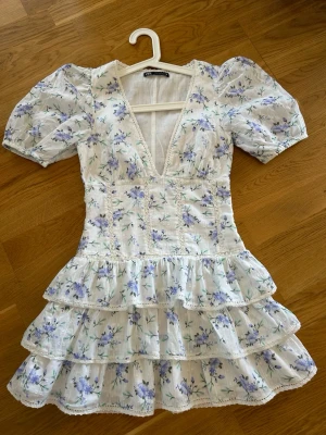  Ny slutsåld student Vit blommig volangklänning från Zara  från Zara - Supersöt kort klänning från Zara i vit bomull med lila och blått blommönster. Djup v-ringning, puffärmar och flera volanger nedtill ger en romantisk vibe. Perfekt för sommarens alla tillfällen!