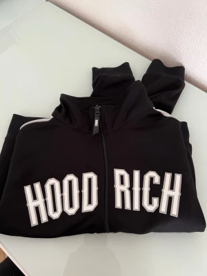 Svart hoodie från Hoodrich med dragkedja - Snygg svart hoodie från Hoodrich med vit text på bröstet och dragkedja framtill. Ärmarna har vita ränder längs sidorna för en sportig look. Tillverkad i mjukt material som känns skönt mot huden. Perfekt för dig som gillar streetwear och vill sticka ut.