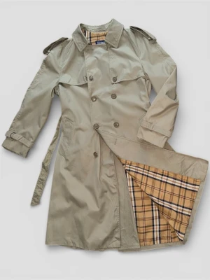 Beige trenchcoat från Burberry - Klassisk beige trenchcoat från Burberry med dubbelknäppning, axelklaffar och bälte i midjan. Insidan har det ikoniska rutiga Burberry-mönstret i beige, svart och rött. Tillverkad i bomullsmix för en tidlös och stilren look.
