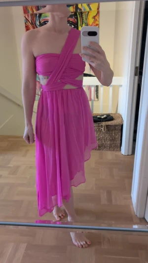 Rosa oneshoulder klänning i polyester - Snygg rosa oneshoulder klänning med asymmetrisk nederkant och omlott-detaljer upptill. Tillverkad i 100% polyester med luftig och lätt känsla. Perfekt för dig som vill sticka ut med en unik och trendig look.