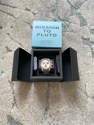 Omega x Swatch Mission to Pluto klocka - Säljer en Omega x Swatch Mission to Pluto från Bioceramic MoonSwatch Collection. Klockan har en beige urtavla med vinröda detaljer och en vinröd bezel. Armbandet är olivgrönt och klockan har kronograf-funktion. Kommer i originalbox med manual.