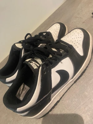 Nike Dunk Low svart/vit sneakers - Säljer ett par Nike Dunk Low sneakers i svart och vitt. Klassisk design med svart swoosh, vita paneler och svarta detaljer. Skorna har lågt skaft, snörning och ovandel i läder. Perfekta för dig som gillar streetwear och vill ha en tidlös sneaker i garderoben. Köpt från Zalando 