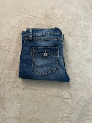 Blå bootcut jeans med fickdetalj - Säljer ett par klassiska blå bootcut jeans med snygg fickdetalj bak och kontrastsömmar. Jeansen har normalhög midja, bälteshällor och knappgylf. Perfekta till en avslappnad stil och passar till det mesta i garderoben.