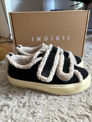 Svarta Inuikii sneakers med fuskpäls -  svarta Inuikii sneakers med beige fuskpälsdetaljer längs kanterna och på kardborrebanden. Skorna har en chunky vit sula och är tillverkade i mockaliknande material. Dom är i storlek 37 och endast använda ett fåtal gånger. Nypris 2400 och köpta på Nelly 
