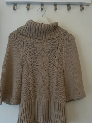 Beige stickad poncho med polokrage - Säljer en beige stickad poncho med bred polokrage och snygga flätstickade detaljer framtill. Ponchon har korta ärmar och bred ribbstickad kant nedtill. Perfekt för lager på lager och riktigt mysig till hösten.