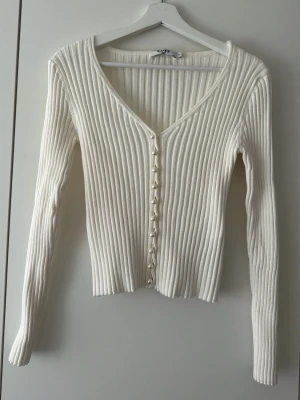 Vit ribbad kofta med pärlknappar - Trendig vit ribbad kofta från NA-KD med långa ärmar och djup V-ringning. Koftan har snygga pärlknappar framtill som ger en elegant touch. Tillverkad i mjukt, stretchigt material som sitter tajt och bekvämt. Perfekt att styla till jeans eller kjol.