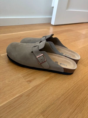 Beige birkenstock boston - Snygga beige tofflor från Birkenstock i mocka med klassisk design och justerbart spänne i metall. Fotbädden är i kork och sulan är svart. Perfekta för dig som gillar stilrena och bekväma skor.