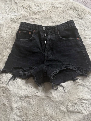 Svarta jeansshorts med råa kanter - Säljer ett par svarta midwaist  jeansshorts med slitna, fransiga kanter och knappar i gylfen. Shortsen har klassiska fem fickor och bälteshällor. Perfekta för en avslappnad och trendig look.