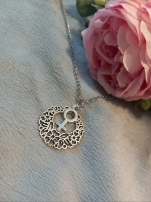 Halsband med kvinnosymbol - Snyggt halsband i silverfärg med ett runt hänge dekorerat med blommönster och en kvinnosymbol i mitten. Perfekt accessoar för att uttrycka girl power och stil. Kedjan är tunn och smidig, och hänget har en modern, detaljrik design.