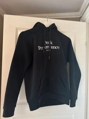 Svart hoodie från Peak Performance - Snygg svart hoodie från Peak Performance med vit logga på bröstet. Klassisk modell med huva och dragsnören samt en stor magficka framtill. Perfekt för en avslappnad och sportig stil. Tillverkad i mjukt material för extra komfort. Betalning via vanlig swish!!