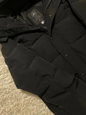 Svart dunjacka från Canada Goose - Snygg svart dunjacka från Canada Goose med klassisk quiltad design och dragkedja samt tryckknappar framtill. Jackan har huva och är fodrad med dun för extra värme. Perfekt för kalla vinterdagar och riktigt stilren look. Det är en liten M storlek 