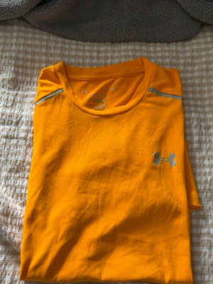 Gul tränings-tshirt från Under Armour - Snygg gul tränings-tshirt från Under Armour med rund halsringning och reflekterande detaljer vid axlarna. Tillverkad i ett lätt och snabbtorkande funktionsmaterial som passar perfekt till träning och sport. Logga på bröstet i silver.