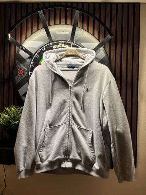 Grå zip hoodie från Polo Ralph Lauren - Klassisk grå hoodie med dragkedja från Polo Ralph Lauren. Tröjan har huva med dragsko, två stora fickor fram och den ikoniska broderade loggan på bröstet. Tillverkad i mjuk bomullsblandning som är skön att ha på sig. Perfekt för en avslappnad och stilren look. Bra skick pris kan diskuteras