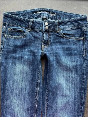 Blå jeans från American Eagle - Snygga blå jeans från American Eagle med klassisk femficksmodell och dubbla knappar i midjan. Jeansen har en bootcut passform och coola slitningar framtill. Tillverkade i mjukt denimtyg som sitter skönt hela dagen.         Kontakta om ni har frågor om mått eller andra saker!💕
