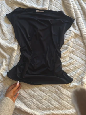 Svart ärmlös topp i syntetmaterial - Säljer en svart, slät och ärmlös topp i mjukt syntetmaterial. Toppen har en enkel och clean design med lätt stretch och smickrande passform. Perfekt att styla med jeans eller kjol för en trendig look.