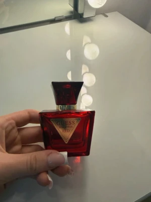 Guess Seductive Red parfym - En elegant parfymflaska från Guess, modell Seductive Red. Flaskan är djup röd med en guldig trekantig etikett framtill och har en matchande röd, fyrkantig kork. Tillverkad i glas med exklusiv känsla och modern design.