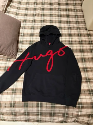 Svart hoodie från Hugo med röd text - Snygg svart hoodie från Hugo med stor röd broderad logga tvärs över bröstet. Klassisk huva och känguruficka framtill. Perfekt för dig som gillar streetwear och vill sticka ut med en fet logga.