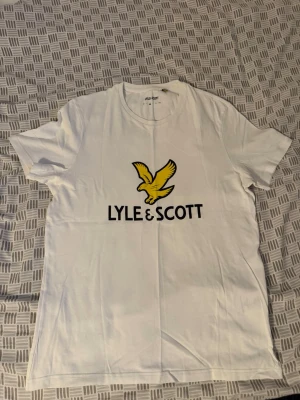 Vit Lyle & Scott t-shirt med tryck - Vit t-shirt från Lyle & Scott med klassiskt tryck av en gul fågel och svart logga på bröstet. T-shirten har rund halsringning och korta ärmar. Perfekt för en clean och stilren look.