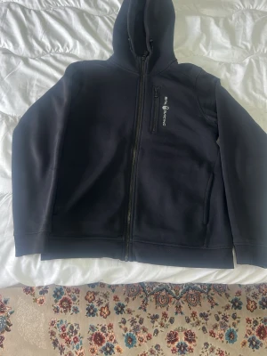 Svart hoodie från Peak Performance - Säljer en svart hoodie från Sail Racing med dragkedja framtill och en bröstficka med logga. Hoodien har två sidofickor och en klassisk huva. Perfekt för dig som gillar stilrena och sportiga plagg. Materialet känns mjukt och bekvämt.