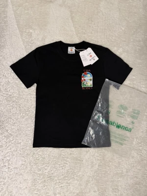 Svart t-shirt från Casablanca - Svart t-shirt från Casablanca med färgglad tryckt grafik framtill som visar ett fönster med blommor och landskap. Klassisk rund hals och korta ärmar. Tillverkad i mjuk bomull för skön känsla. Perfekt för dig som gillar stilrena plagg med unik detalj. Storlek S men passar mer som XS 👍Priset kan diskuteras 