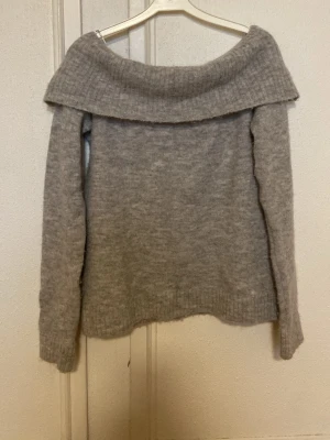 Grå off shoulder stickad tröja Gina Tricot - Mysig grå stickad tröja från Gina Tricot med bred off shoulder-krage och långa ärmar. Tröjan har en mjuk och fluffig känsla, perfekt för kyliga dagar. Ribbstickade muddar vid ärmslut och nederkant ger en snygg finish.