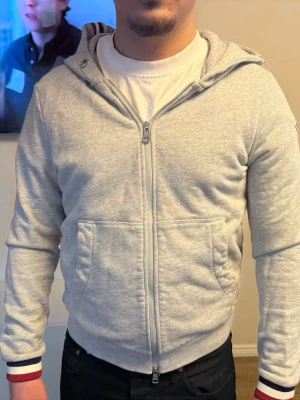 Ljusgrå hoodie med dragkedja - Snygg ljusgrå hoodie med dragkedja framtill och två stora fickor. Huva med dragsko och ribbade muddar med röd, vit och marinblå detalj på ärmsluten. Tillverkad i mjukt sweatshirtmaterial, perfekt för en avslappnad stil.