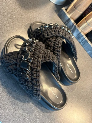 Helt nya inuikii med pärlor - Säljer ett par svarta sandaler från Inuikii med breda, stickade band dekorerade med stora svarta pärlor och stenar. Sulan är svart och har en bekväm formad fotbädd. Perfekta för dig som vill ha en unik och trendig look.