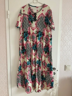 Mönstrad långklänning med rosor - Unik långklänning med kort ärm i cremevitt tyg, dekorerad med stora rosa och turkosa rosor samt svartvita grafiska mönster. Rund halsringning med dekorativa knappar och pärldetaljer. Luftigt material, perfekt för sommarens alla tillfällen.