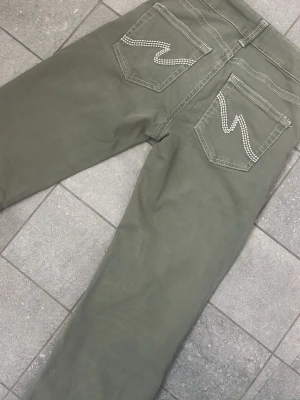 Gröna bootcut jeans från Nelly - Snygga gröna bootcut jeans från Nelly med vita kontrastsömmar och dekorativa detaljer på bakfickorna. Jeansen har normalhög till låg midja, klassisk femficksdesign och stängs med dragkedja och dubbla knappar. Perfekta för en trendig och avslappnad look. Små slitningar pga användning längst ner på benet, men inget direkt synligt på. Storlek 34! 