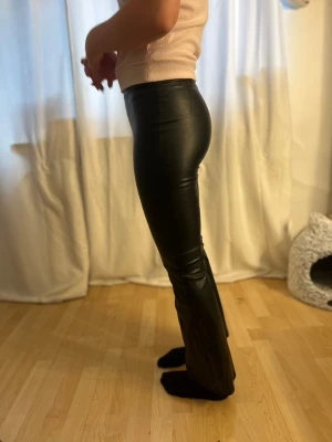 Svarta fuskskinnbyxor med slits - Snygga svarta byxor i fuskskinn med tight passform och slits nertill på benen. Byxorna har utsvängda ben, vilket ger en trendig siluett. matchar lätt med olika toppar. Fuskskinnet har spruckit på höften och lite mellan benen, som man kan se på bilderna. Men då det är tyg under ”skinnet” så ser man inte igenom, trots sprickorna. Och sprickan mellan benen syns inte alls när man har på sig dom. passar längd mässigt på dig som är 157 - 162 cm lång 