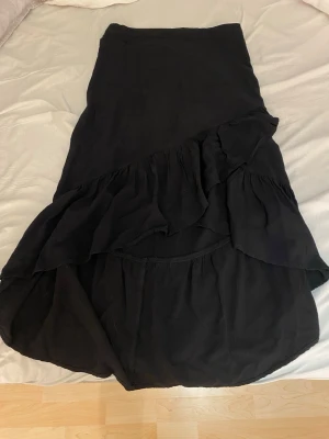 Svart långkjol med volang - Säljer en svart långkjol med volangdetaljer och asymmetrisk nederkant. Kjolen har en mjuk och flowy känsla, perfekt för dig som gillar en enkel men ändå trendig stil. Resår i midjan för skön passform.