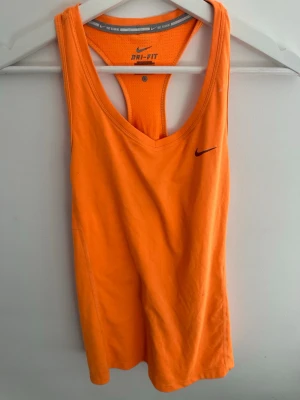 Orange träningslinne från Nike - Snyggt orange träningslinne från Nike med Dri-FIT-material som andas. Linnet har brottarrygg och en liten svart Nike-logga på bröstet. Perfekt för gymmet eller löprundan när du vill ha något lätt och bekvämt.