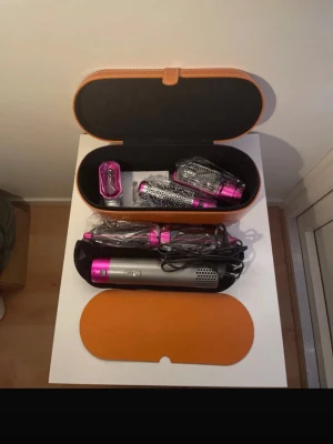 Dyson Airwrap med tillbehör och väska - Säljer en Dyson Airwrap i silver och rosa med flera stylingtillbehör som borstar och locktillbehör. Allt kommer i en snygg brun förvaringsväska med svart insida. Perfekt för dig som vill fixa håret på olika sätt med ett enda verktyg.