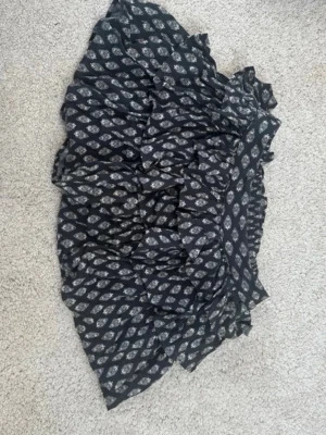 Svart volangkjol från Divided - Kort svart kjol från Divided med vita blommönster och flera volanglager. Kjolen har resår i midjan för skön passform och är tillverkad i polyester med bomullsfoder. Perfekt för en trendig och avslappnad look.