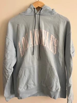 Ljusblå hoodie med Los Angeles-tryck - Snygg ljusblå hoodie med stor ficka framtill och huva med svarta snören. På bröstet står det 'LOS ANGELES' i stora vita bokstäver. Perfekt för en chill och avslappnad stil.