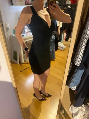 Svart omlottklänning med v-ringning - Snygg svart kortklänning från Filippa K med djup v-ringning. Klänningen är ärmlös och har en figurnära passform som framhäver siluetten. Perfekt för dig som gillar en stilren och klassisk look.