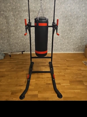 Svart och röd boxningsställning - Säljer en stabil boxningsställning med svart och röd boxsäck. Ställningen har flera grepphandtag med röda detaljer och är tillverkad i metall. Perfekt för hemmaträning inom boxning eller styrka. Boxsäcken hänger i kedja och har matchar. Med denna köpet får du med dig boxningsäcken och även boxning handskar. Tyvärr går det inte att frakta den, köparen måste hämta. 