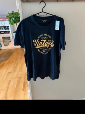 Svart t-shirt med vintage-tryck - Snygg marinblå t-shirt från Livergy med stort gult vintage-inspirerat tryck på bröstet. Rund halsringning och korta ärmar. Tillverkad i mjuk bomull för skön känsla. Perfekt för dig som gillar retrostil och grafiska prints.